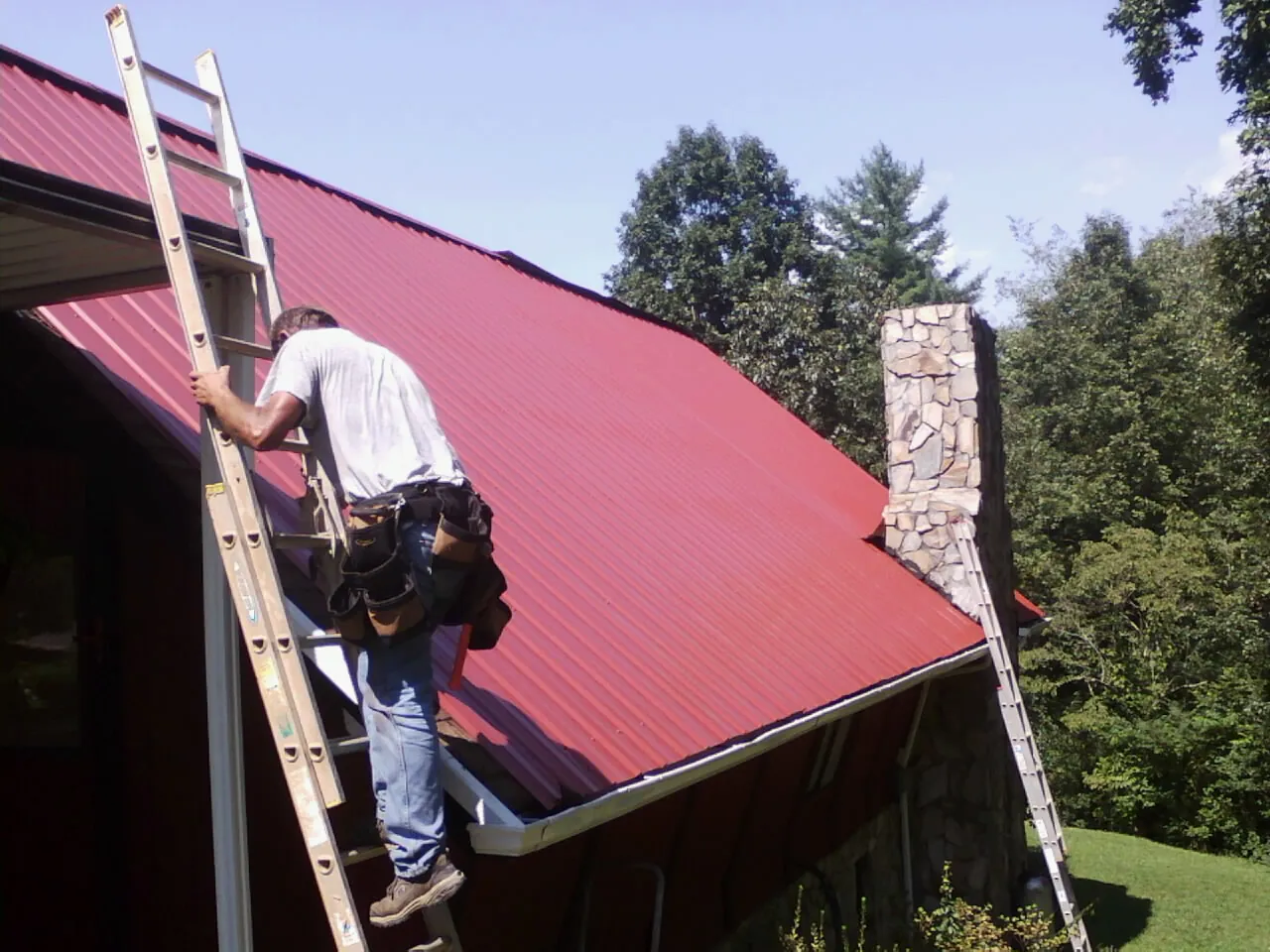 Roofer on a ladder beside a red metal roof and stone chimney — Soffit & Fascia Repair by Roofing Brothers in Makoti